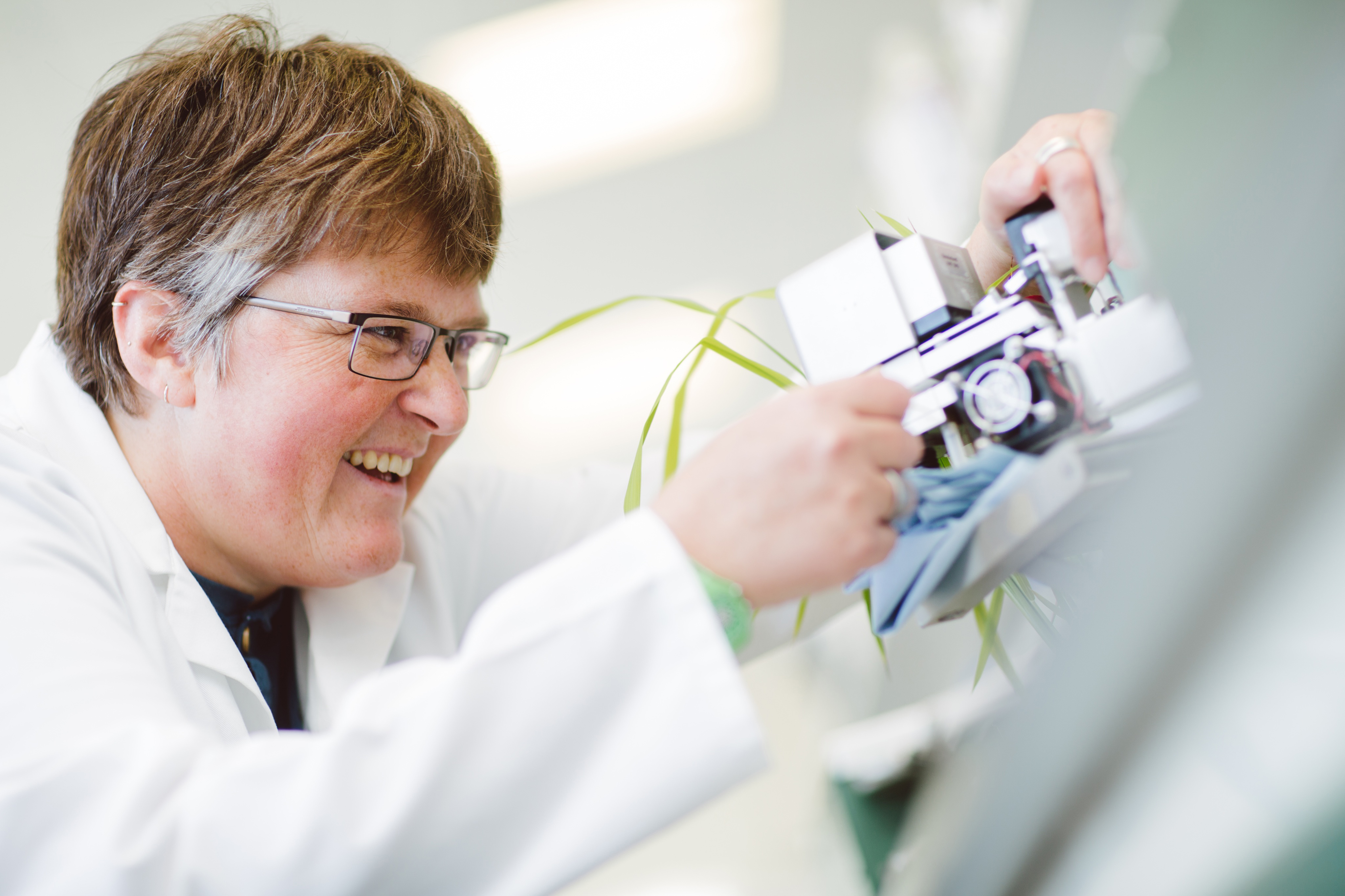 A woman in a lab coat smiles while taking a measurement of leaves with a device.
