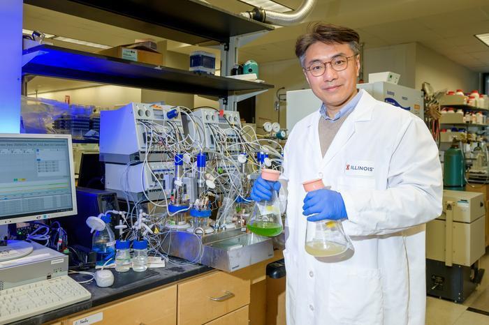 A man in a lab setting in a white lab jacket holds two large beakers with low levels of liquid. One liquid is green and the other is yellow.