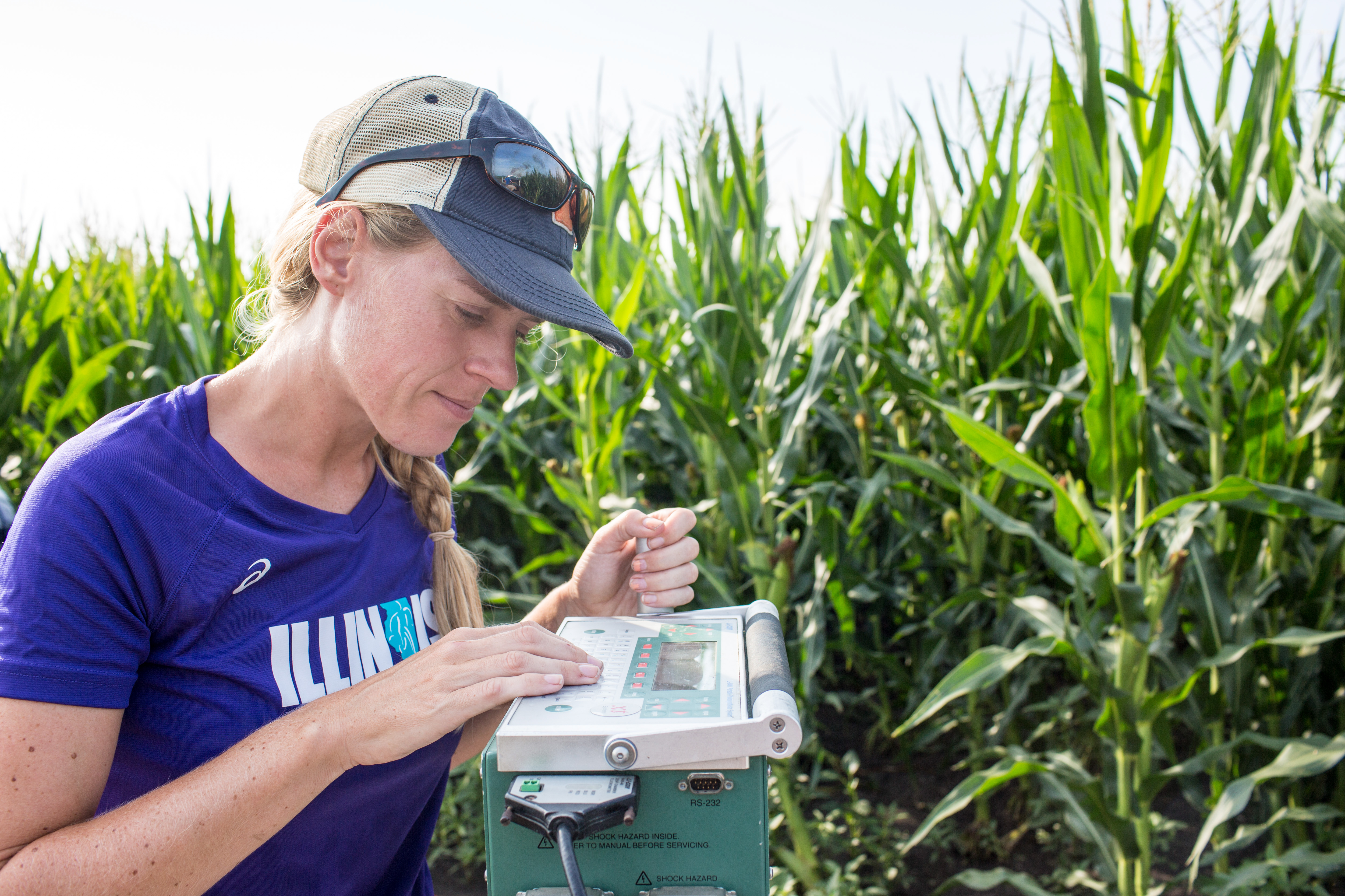 Katherine Meacham takes a reading on a metal-box structure with a corn field in the background.