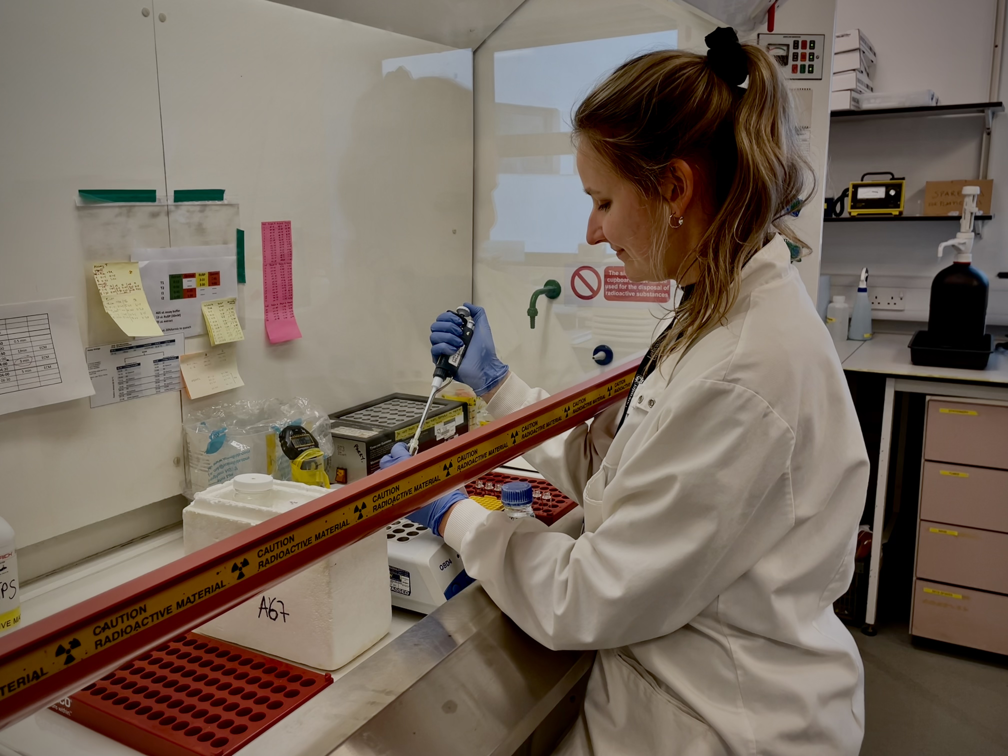 A woman in a lab space works with her arms under a protective bar surrounded by standard lab equipment.