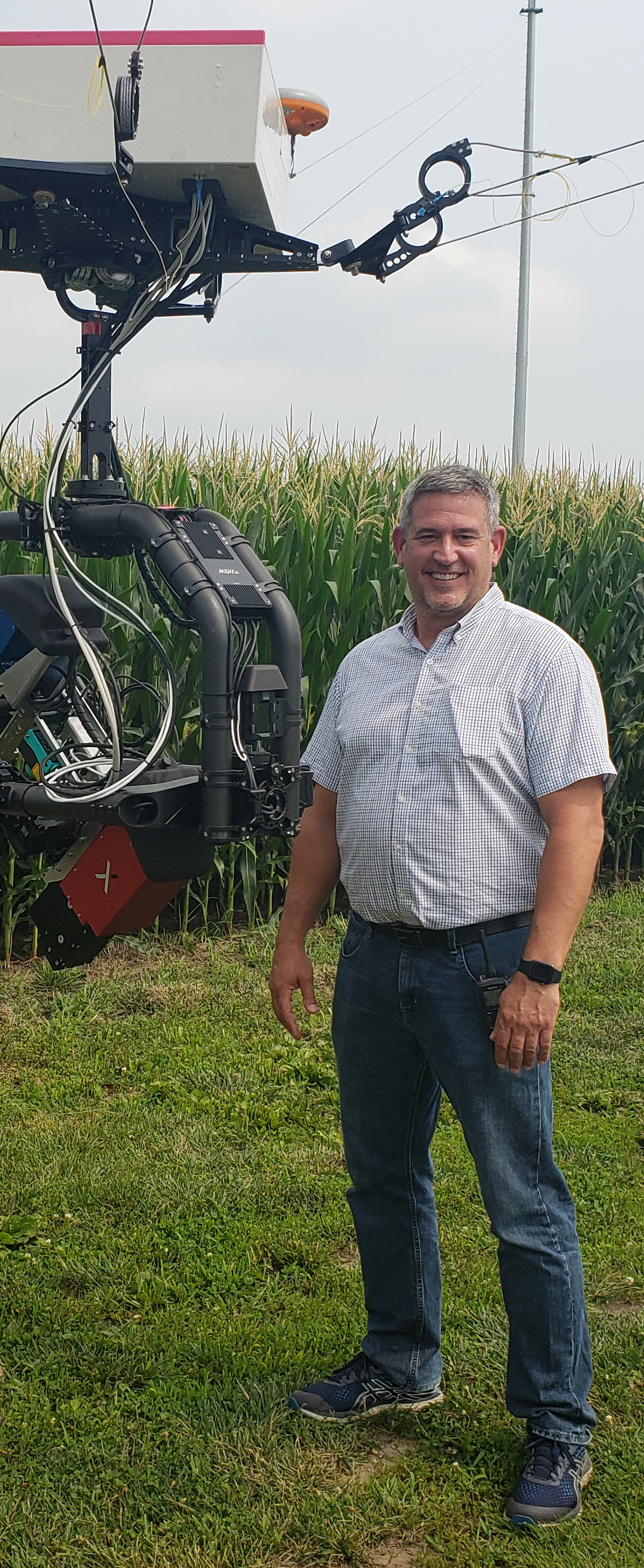 Carl Bernacchi standing next to a camera at torso level in a field with corn in the background.