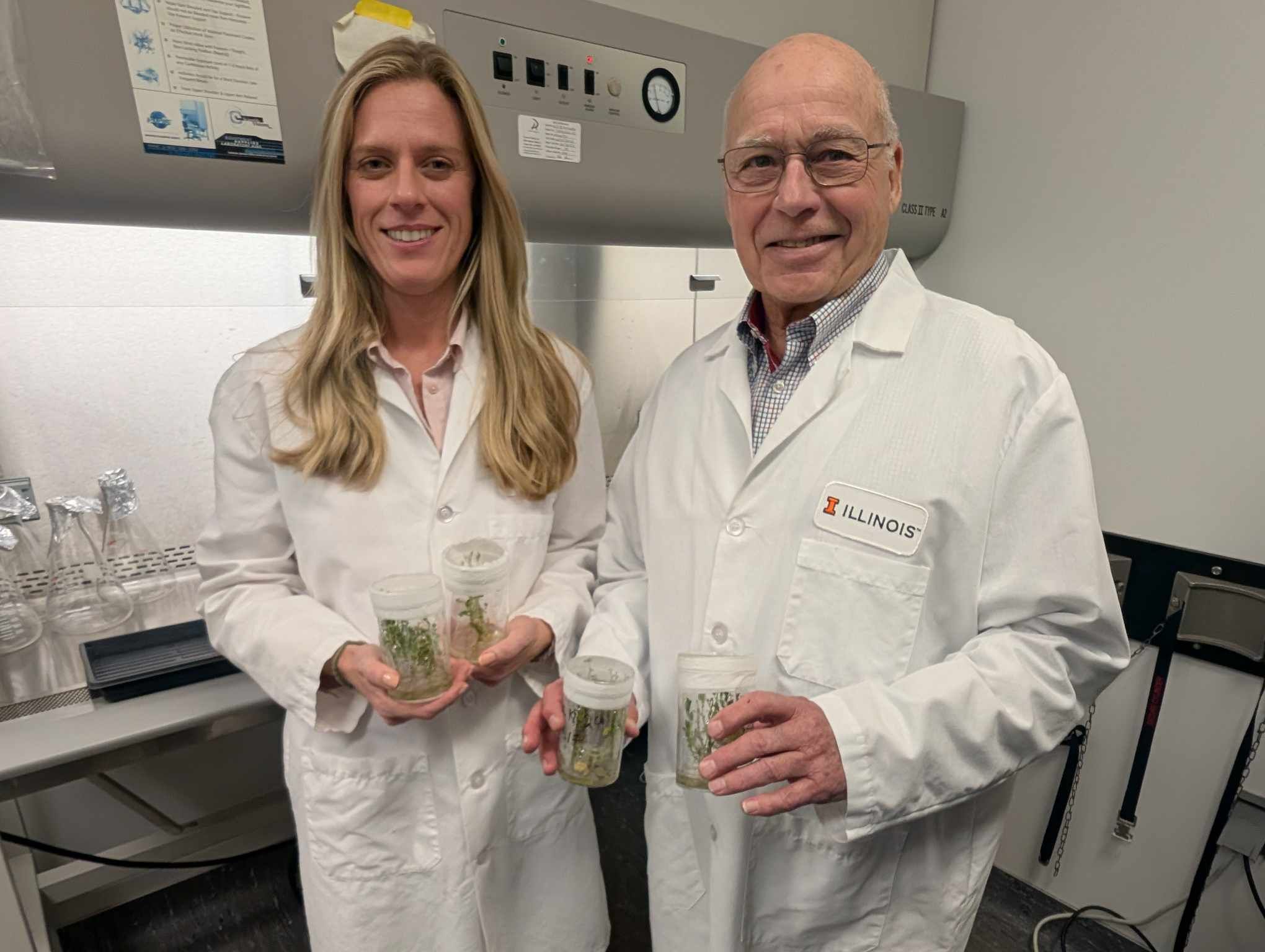 A woman and a man in white lab coats hold containers with young green plants in a lab setting.