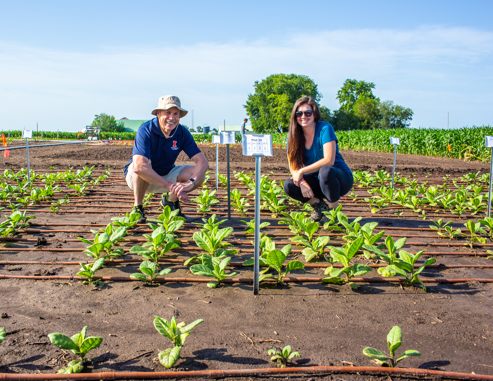 A man and a woman smile at the camera while standing among rows of young, green plants.