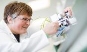 A woman in a lab coat smiles while taking a measurement of leaves with a device.