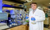 A man in a lab setting in a white lab jacket holds two large beakers with low levels of liquid. One liquid is green and the other is yellow.