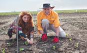 A red headed woman on the left and a man in a hat on the right work in a field with brown dirt.